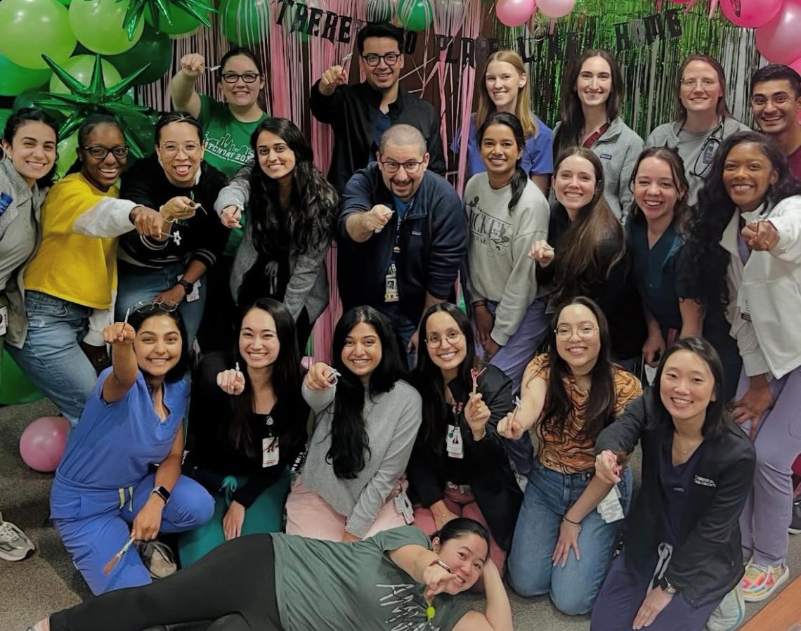 A large group of smiling students pointing at the camera, surrounded by pink and green party decorations.