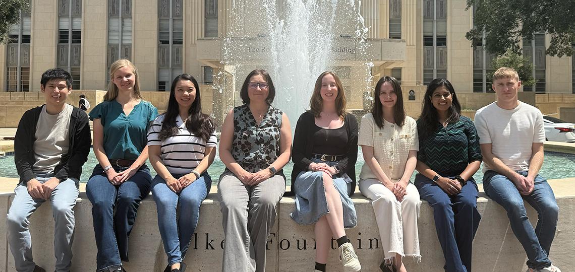 A group of eight people sitting on the edge of a fountain in front of a large stone building with columns and windows, under bright daylight.