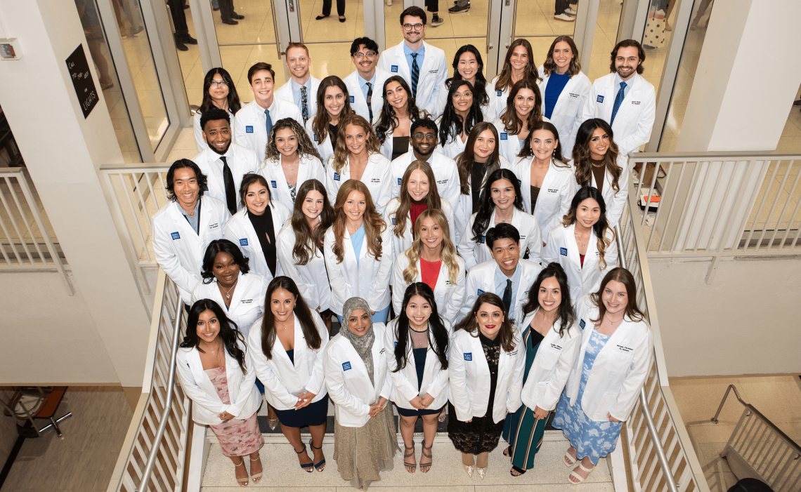 A large group of students wearing white coats posing on a staircase, as seen from above.