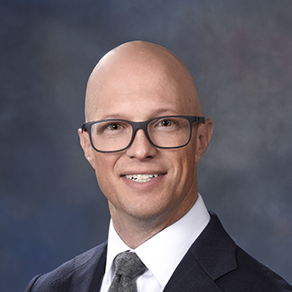 A professional headshot of a man wearing black rectangular glasses, a dark suit jacket, a white dress shirt, and a gray tie. He is smiling slightly and posed in front of a dark, blurred background.
