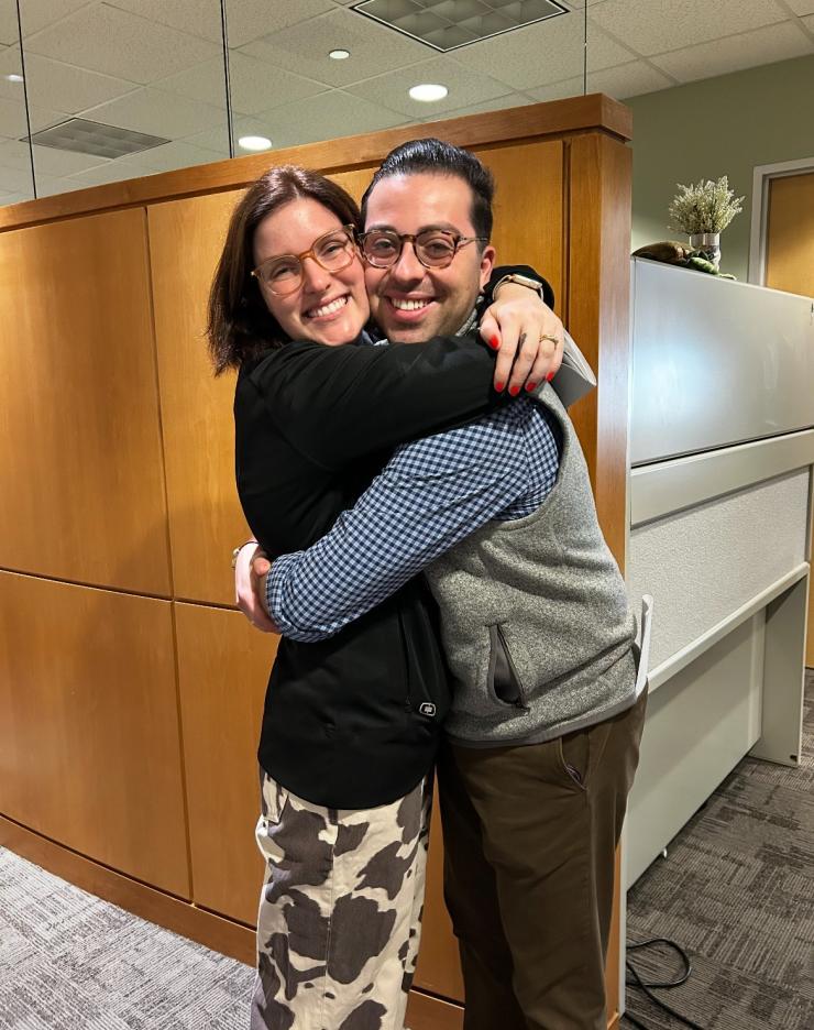 Two people hugging in an office, with wood paneling and office furniture in the background.