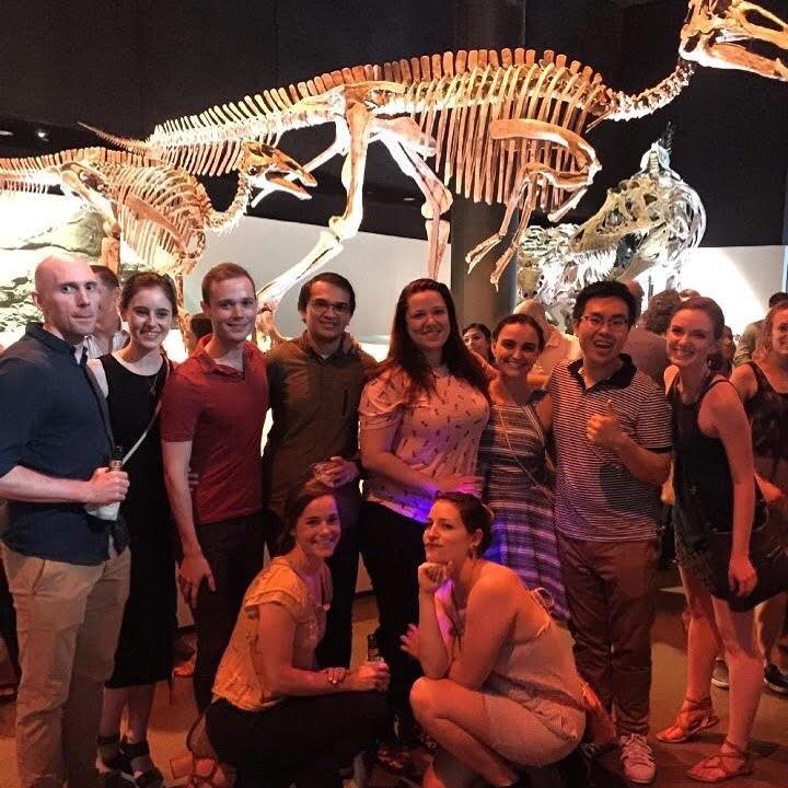 A group of students in a museum posing in front of a large dinosaur skeleton
