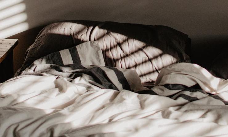 An unmade bed with a sunbeam from an open window shining onto a pillow.