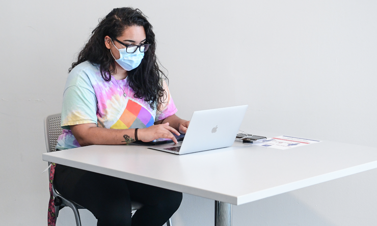 A student in a mask sits at a desk studying on their laptop.