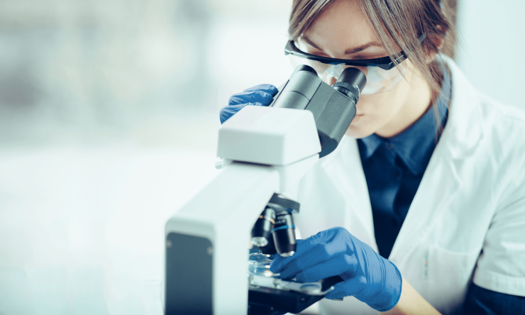 A researcher wearing glasses and protective gloves looks at a slide under a microscope.