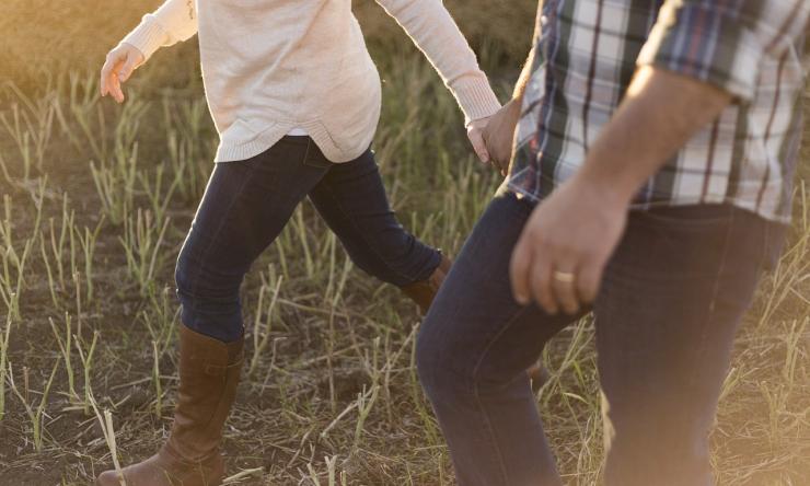 Photo of a couple walking hand in hand through a grassy area only showing couple from the chest down. 