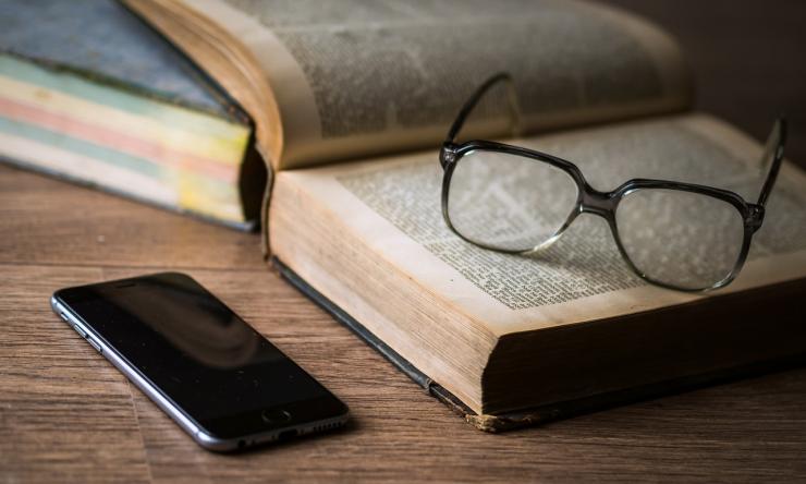 Photo of an open book with reading glasses sitting on the open pages and a cell phone next to the book. 