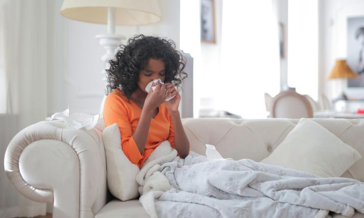 A woman sits on her couch sneezing and covered in a blanket