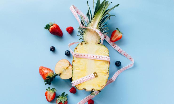 Photo of a pineapple sliced in half surrounded by berries all wrapped in a measuring tape to symbolize healthy eating and weight loss. 