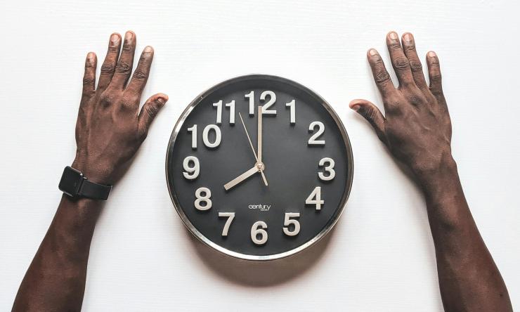 Photo taken from above of a clock face up on a table with a man's hands resting on either side of the clock. 