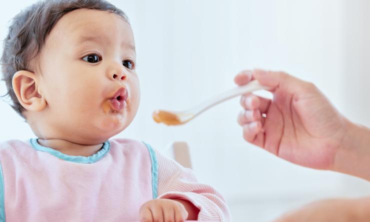 Baby eating solid baby food from a spoon being held by caretaker.