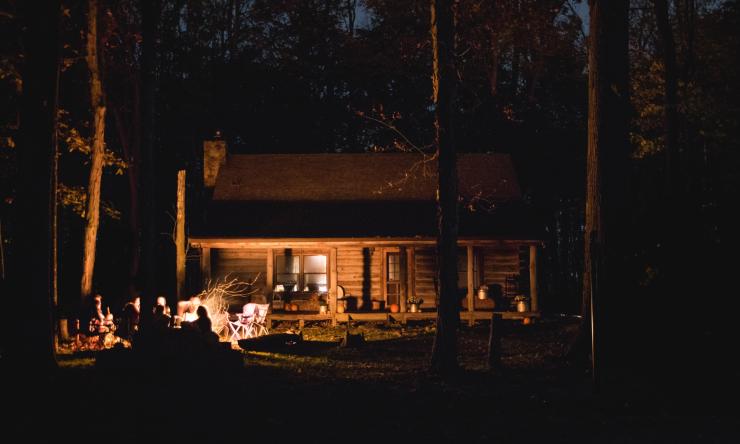The image is a photo taken at night of a cabin in the woods, a group of people are sitting around a camp fire in front of the cabin.