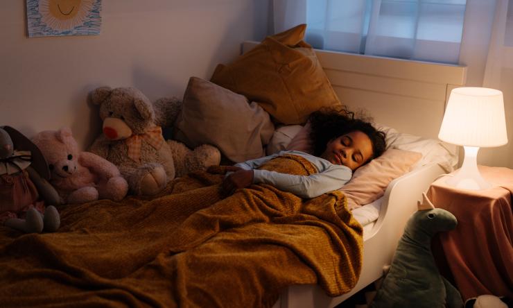 Young child sleeping in her bed with lamp on. Surrounded by stuffed animals.