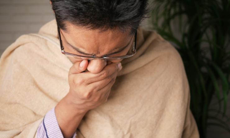 Photo of a man coughing, covering his mouth. He is wrapped in a blanket to indicate he isn't feeling well. 