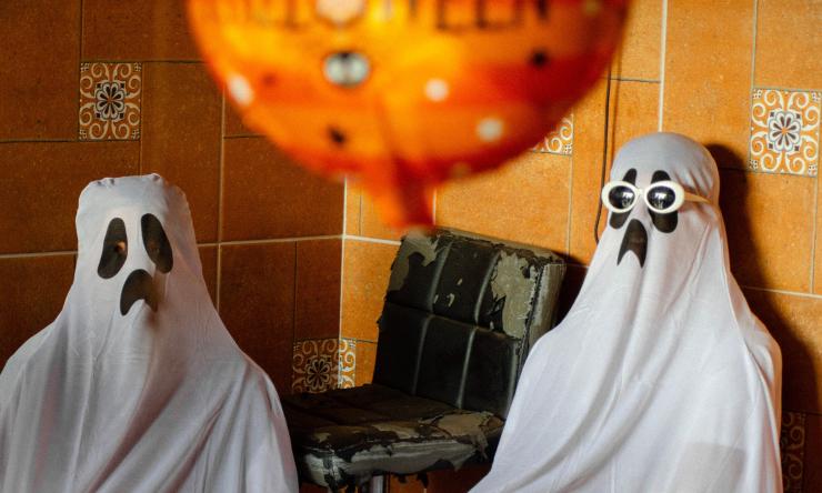 Two people wearing white sheet ghost costumes sitting in a corner of a room. 